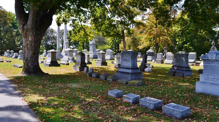 A photo of various graves in lines at the historical site: Glen Cove Cemetery. Trees are pictured, with orange, red, and brown leaves. It's a sunny day.